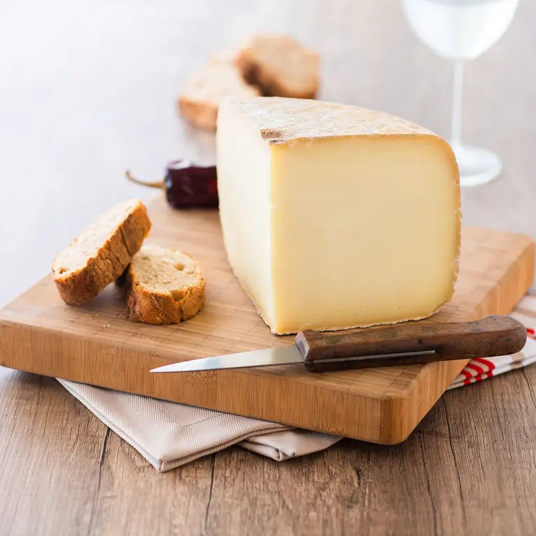 Quart de meule de fromage basque à pâte pressée, posé sur planche à découper en bois avec couteau à manche en bois et tranches de pain rustique; ambiance chaleureuse, lumière douce, fond de table en bois, pour un catalogue, d'un artisan fromager.