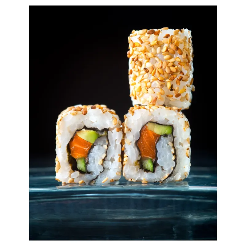 Maki sushi uramaki au saumon et avocat, enrobé de graines de sésame doré, présenté en gros plan sur fond noir, éclairage studio mettant en valeur la texture du riz et la fraîcheur—photo produit professionnelle pour boutique en ligne.