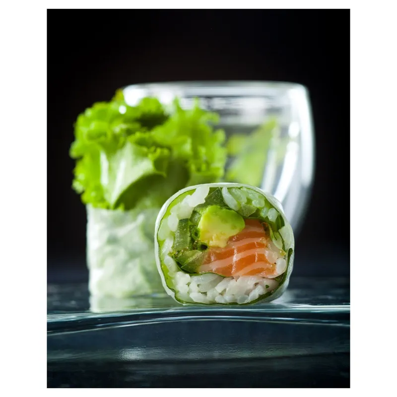 Rouleau de printemps / sushi roll transparent garni de saumon cru, avocat, salade et riz, tranche en gros plan sur fond noir, éclairage studio soulignant la fraîcheur et les textures—packshot culinaire professionnel.