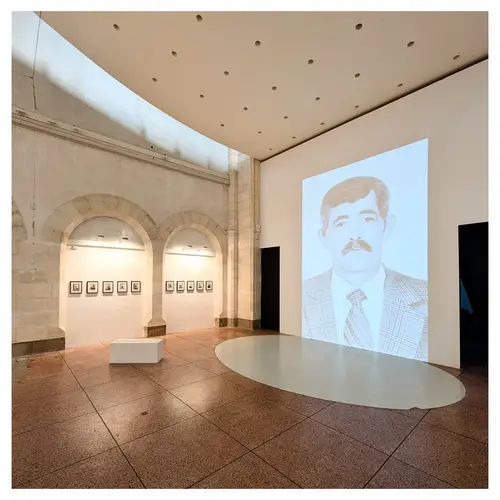 Vue d’ensemble d’une exposition dans une ancienne nef avec projection murale d’un portrait, séries de petits tirages encadrés sous arcades et dispositif scénographique au sol.