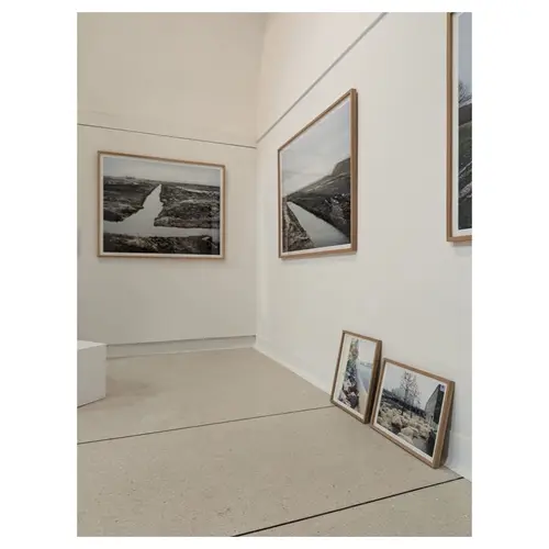 Mur de galerie avec grands tirages de paysage encadrés et deux cadres posés au sol en attente d’accrochage, scénographie en cours.