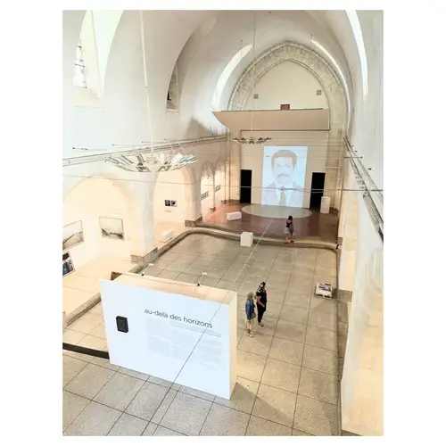 Perspective depuis la mezzanine sur l’ensemble de l’exposition : nef voûtée, cimaises centrales, projection d’un portrait au fond et visiteurs en parcours.