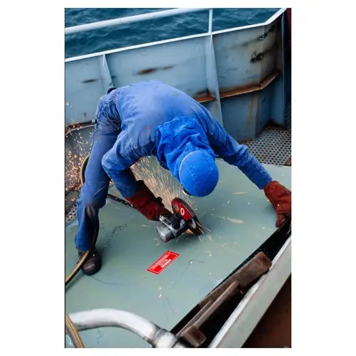 Ouvrier en combinaison bleue utilisant meuleuse avec gerbe d'étincelles sur pont de navire, chantier naval