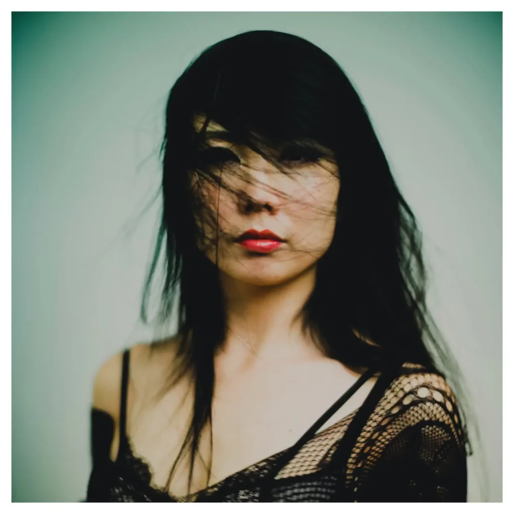 Portrait couleur artistique d’une femme aux longs cheveux noirs balayés par le vent, plan poitrine, regard caméra, lèvres rouges, expression mystérieuse, top en dentelle noire à bretelles glissant sur l’épaule, lumière douce, fond studio vert-gris