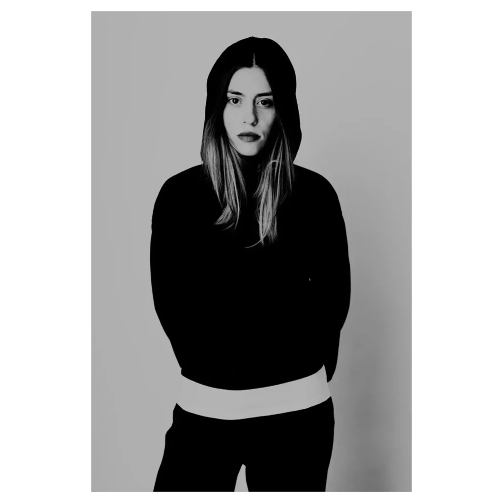 Portrait mode en noir et blanc d’une femme portant un sweat à capuche noir, plan américain, regard caméra, expression sérieuse, cheveux longs sortant de la capuche, t-shirt blanc visible à la taille, pantalon sombre, éclairage contrasté, fond studio gris