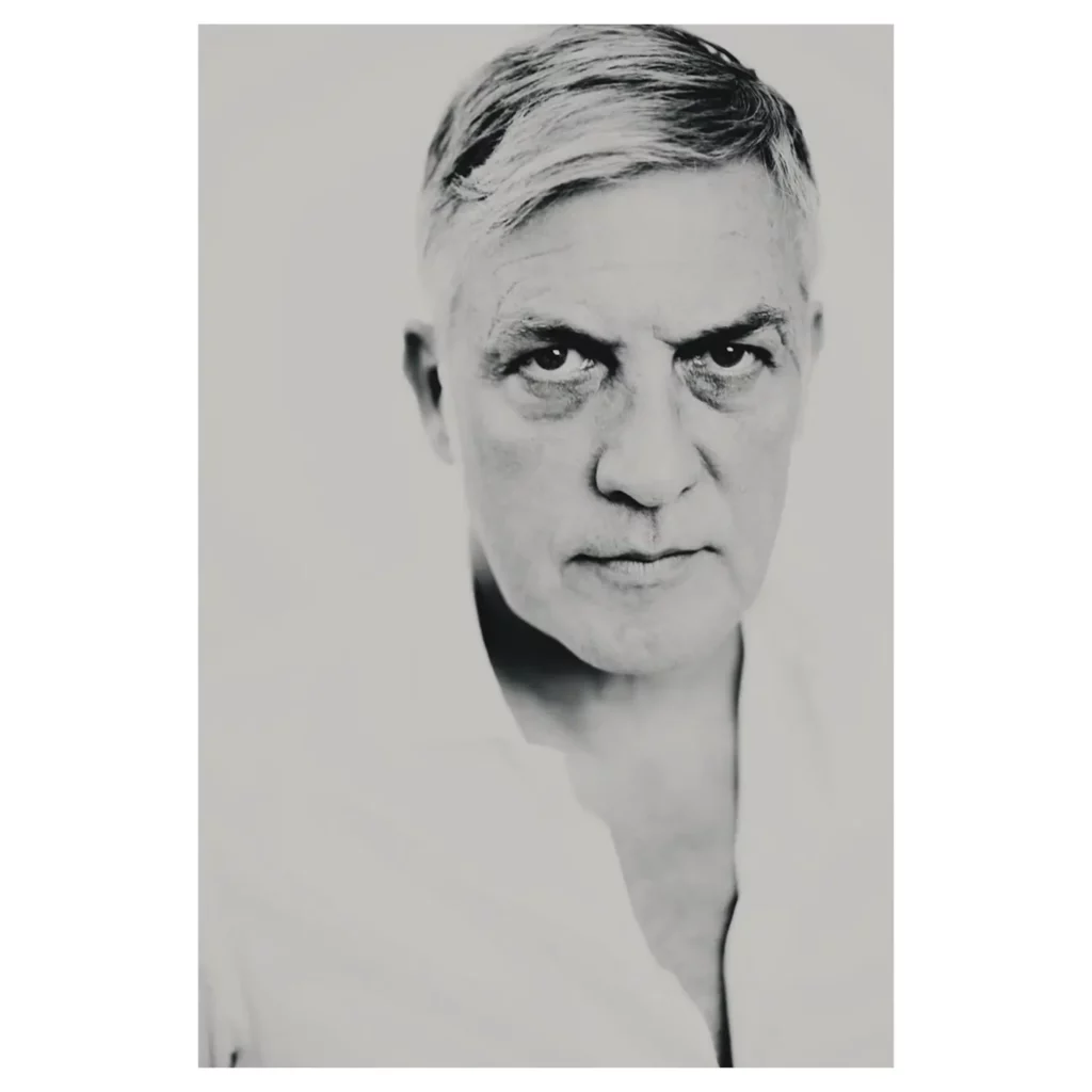 Portrait professionnel noir et blanc d’un homme mature aux cheveux gris courts, gros plan poitrine, regard intense vers l’objectif, éclairage haute clé, chemise blanche col ouvert, fond studio clair.