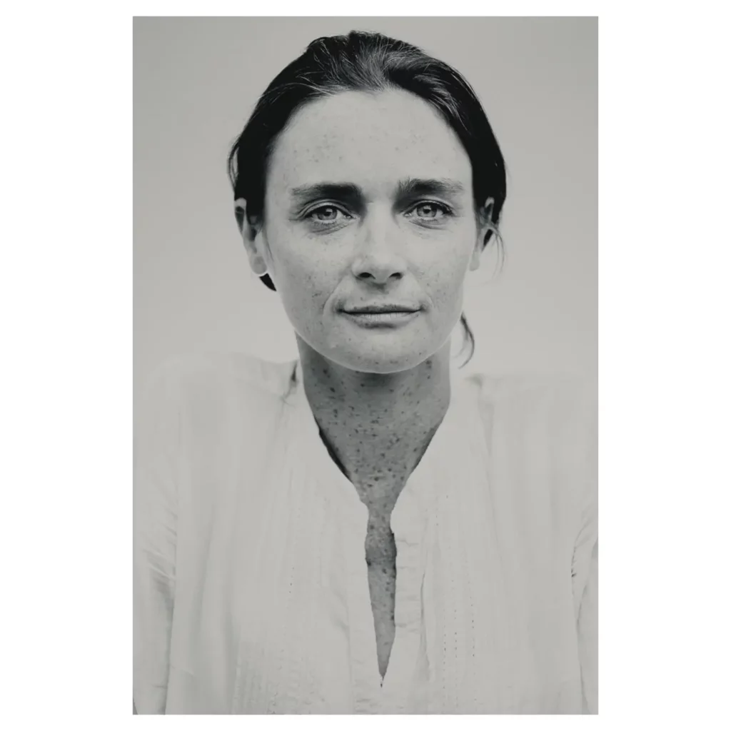 Portrait noir et blanc d’une femme aux taches de rousseur, cheveux relevés, plan poitrine, regard caméra, lumière douce, chemise blanche, fond studio neutre — photographe de portrait à Bordeaux.