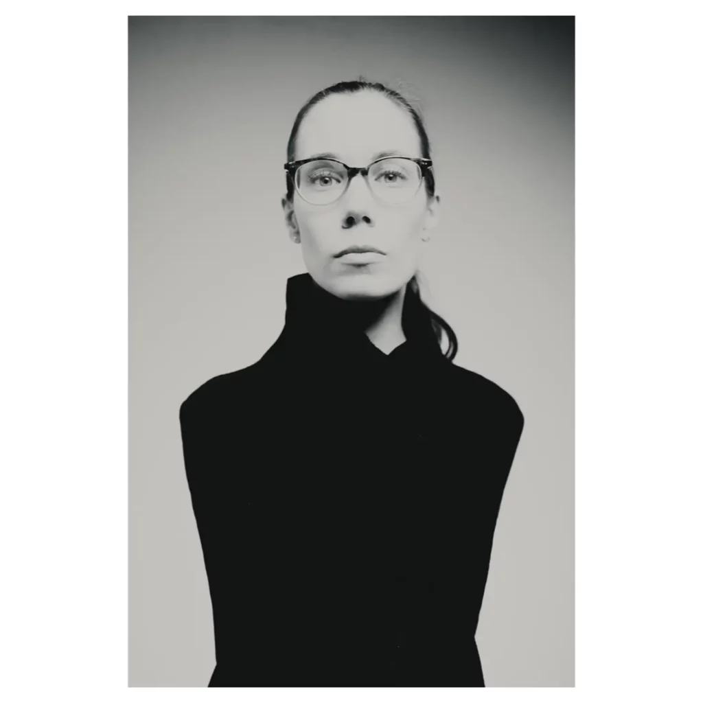 Portrait professionnel noir et blanc d’une femme portant des lunettes, cheveux tirés en queue-de-cheval, plan mi-corps, expression sérieuse, col montant noir minimaliste, éclairage studio doux avec vignettage, fond dégradé gris