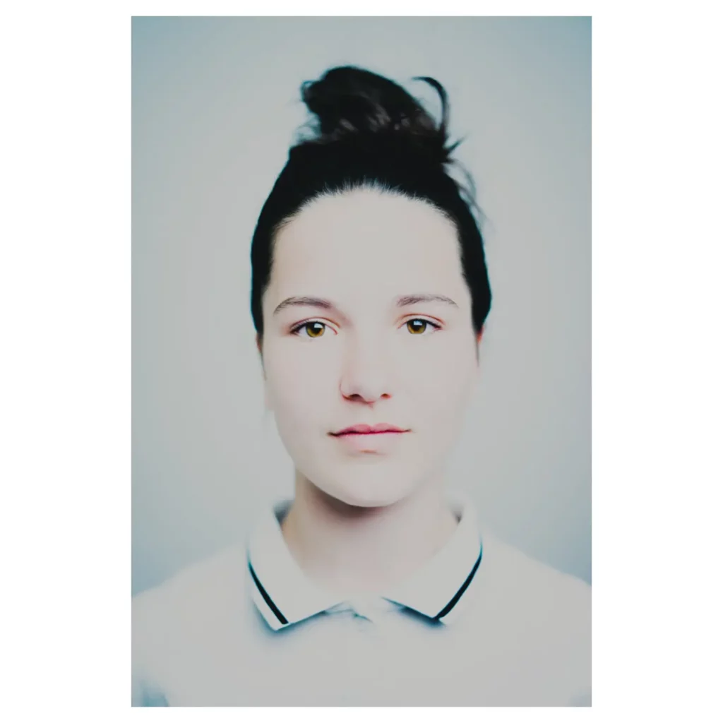 Portrait couleur en haute clé d’une jeune femme, chignon, plan poitrine, regard caméra, peau lumineuse, polo blanc à liserés, fond studio gris clair