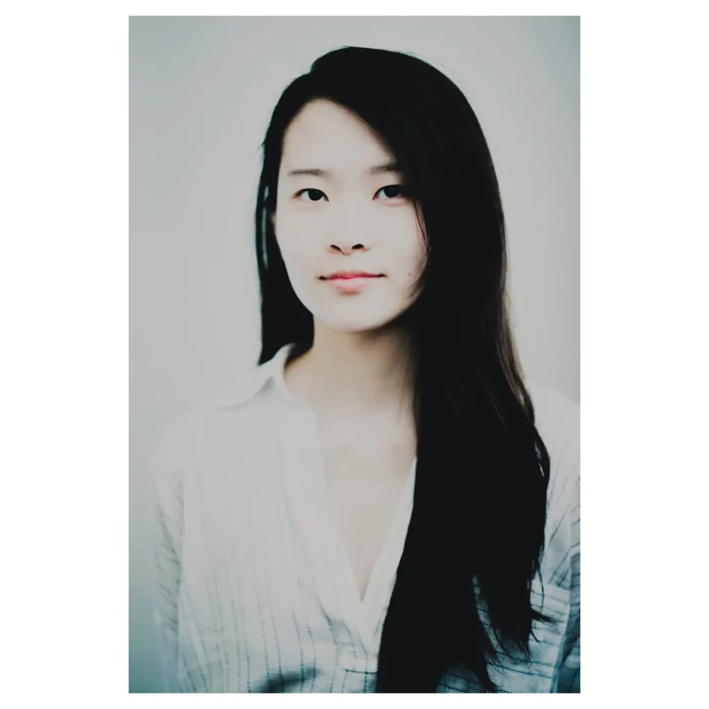 Portrait artistique couleur en haute clé d’une jeune femme aux longs cheveux noirs portés sur une épaule, plan poitrine, léger sourire, regard caméra, lumière douce, chemise blanche à fines rayures, fond studio gris clair