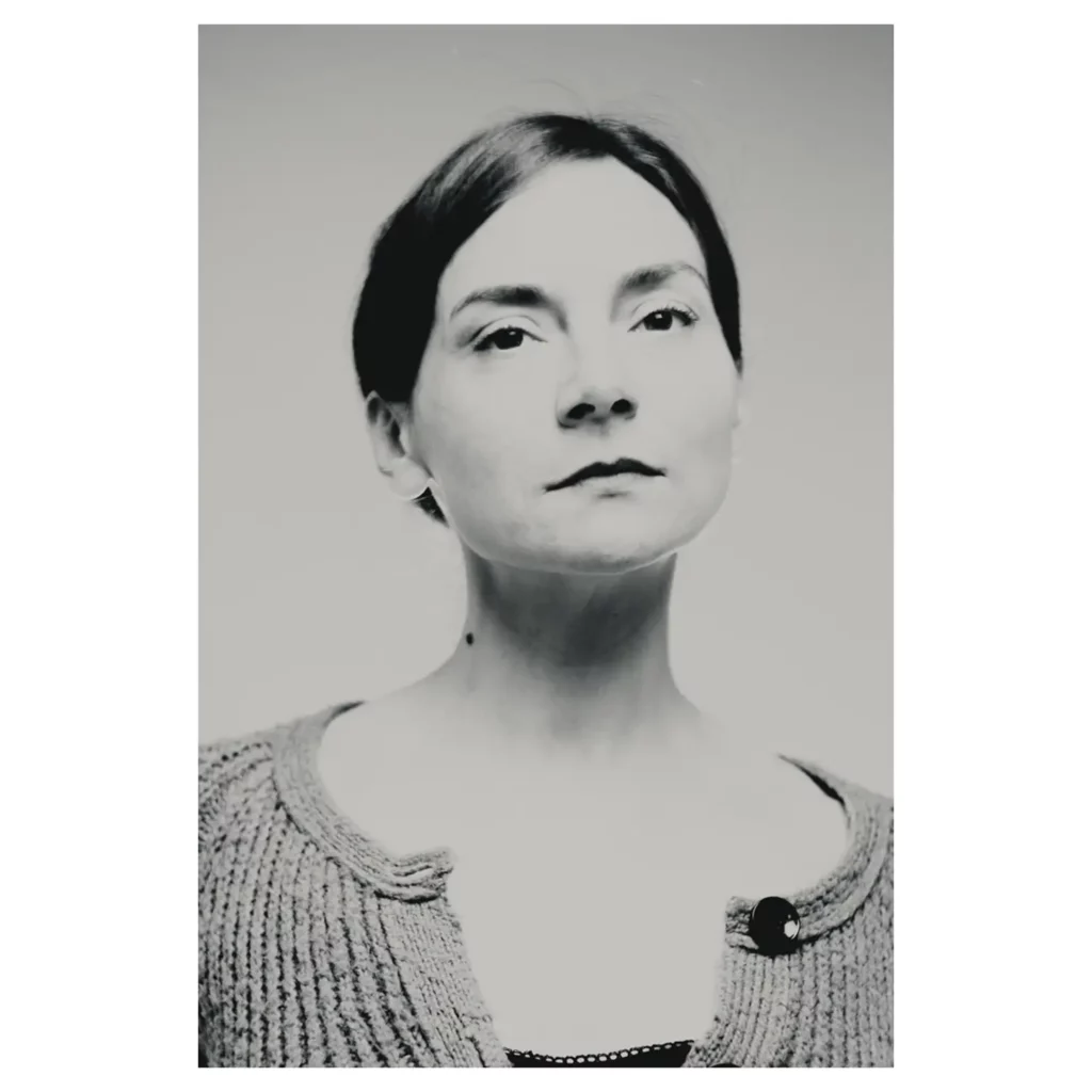 Portrait noir et blanc d’une femme aux cheveux bruns relevés, plan poitrine, menton légèrement relevé, expression assurée, lumière haute clé, cardigan en maille à gros bouton sur débardeur sombre, fond studio photo gris