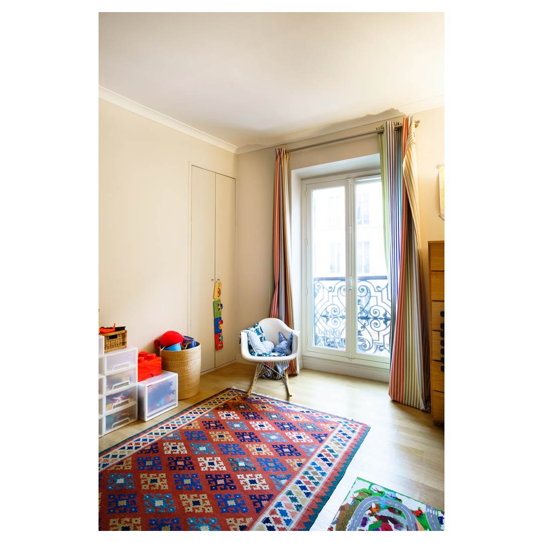 Chambre enfant lumineuse, tapis kilim rouge, rideaux rayés, balcon en ferronnerie — photo d’architecture intérieure à Paris.