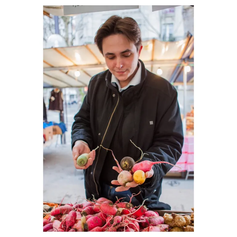 Portrait d’un chef culinaire sur un marché tenant des radis multicolores (rose, noir, jaune) au-dessus d’un étal garni ; ambiance de stand, bokeh de lampes, mise au point sur la fraîcheur des légumes—photographie professionnelle pour site e-commerce / branding alimentaire.