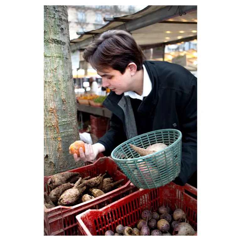 Jeune chef de cuisine, sélectionnant des légumes racines dans des cagettes rouges au marché, panier vert à la main (betteraves, racines variées), scène de stand en extérieur, lumière naturelle—photographie professionnelle pour site e-commerce / storytelling de marque alimentaire.