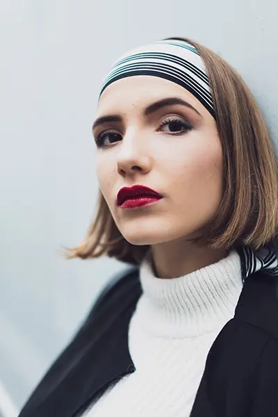 Portrait mode en lumière naturelle d’une femme au carré plongeant, bandeau rayé et rouge à lèvres, sur fond bleu clair — photographe portrait à Bordeaux.