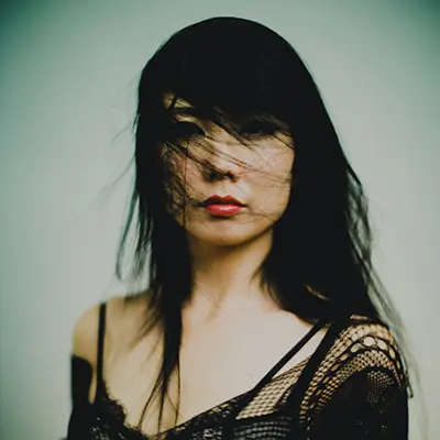 Portrait studio éditorial d’une femme aux cheveux noirs, mèches devant le visage et rouge à lèvres, tenue noire en résille, fond clair — photographe portrait à Bordeaux.