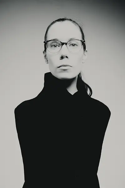 Portrait studio noir et blanc d’une femme portant des lunettes, cadrage buste, col montant, fond gris clair — photographe portrait à Bordeaux.