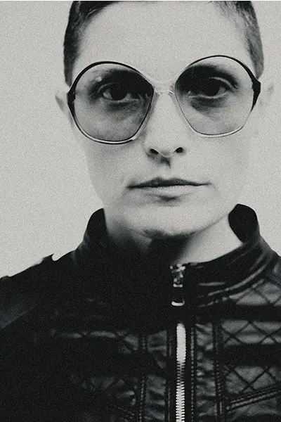 Portrait studio noir et blanc d’une femme avec lunettes rondes, cadrage serré, veste en cuir zippée, rendu grain argentique — photographe portrait à Bordeaux.