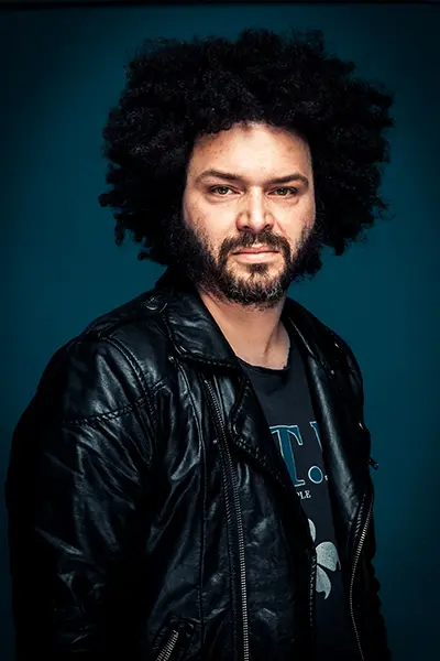 Portrait studio d’un homme avec cheveux afro et barbe, veste en cuir, cadrage buste sur fond bleu, lumière douce — photographe portrait Bordeaux Paris.