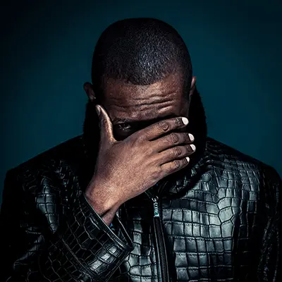 Portrait studio d’un homme sur fond bleu, main devant le visage laissant apparaître le regard, veste en cuir texturée, ambiance dramatique — photographe portrait Bordeaux Paris.