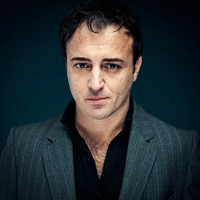 Portrait studio d’un homme en veste à fines rayures et chemise noire sur fond bleu, cadrage buste, regard caméra, rendu professionnel — photographe portrait Bordeaux Paris.