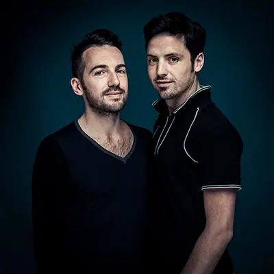 Portrait studio d’un duo de deux hommes sur fond bleu, cadrage mi-buste, regard caméra, lumière douce et contraste maîtrisé — photographe portrait Bordeaux Paris.