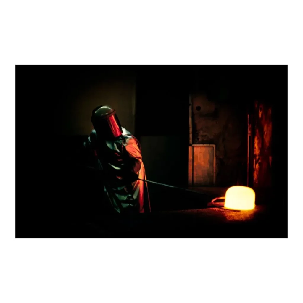 Photographie de reportage industriel montrant un artisan en équipement de sécurité manipulant un métal en fusion rougeoyant dans une fonderie.