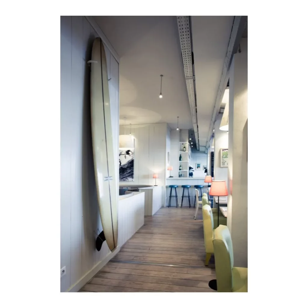 Vue en enfilade d'un restaurant moderne au style surf avec planche accrochée au mur, parquet clair et espace bar.
