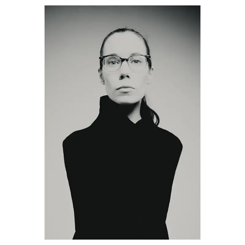 Portrait professionnel noir et blanc d’une femme portant des lunettes, cheveux tirés en queue-de-cheval, plan mi-corps, expression sérieuse, col montant noir minimaliste, éclairage studio doux.