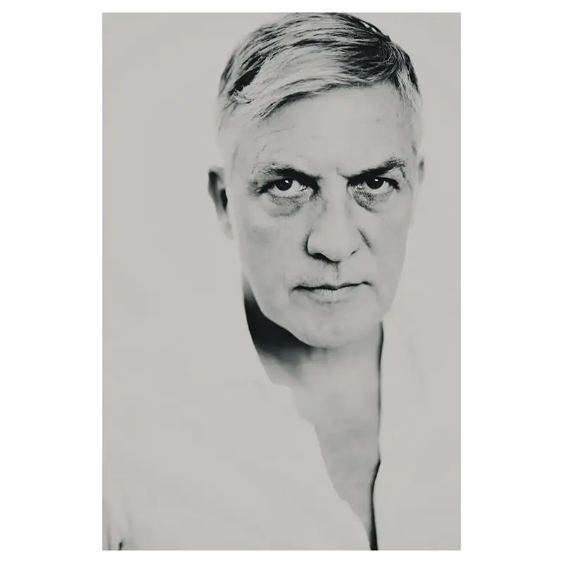 Portrait photographe professionnel, en noir et blanc, d’un homme mature aux cheveux gris courts, gros plan poitrine, regard intense vers l’objectif, chemise blanche col ouvert.