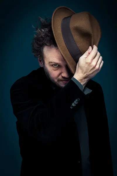 Portrait cinématographique en studio d'un homme soulevant un chapeau de feutre marron sur fond bleu.