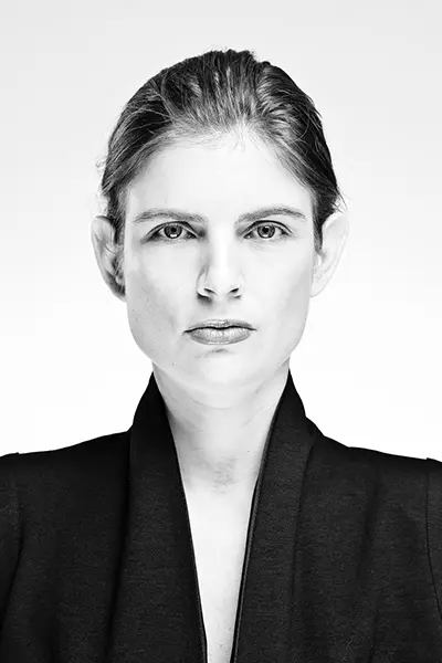 Portrait éditorial en noir et blanc high key d'une femme en veste noire ajustée sur fond blanc.