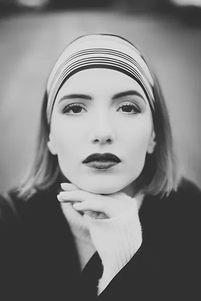 Portrait en noir et blanc d'une femme aux cheveux courts, portant un bandeau rayé. Lumière douce et fond flou confèrent une profondeur graphique. Regard frontal intense, lèvres sombres, main délicatement posée sous le menton. Atmosphère épurée et introspective.
