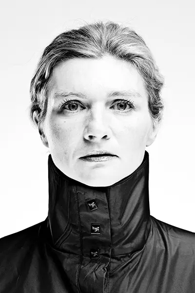 Portrait studio en noir et blanc high key d'une femme portant une veste sombre à haut col structuré, sur fond blanc.