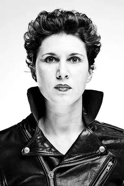 Portrait studio en noir et blanc high key d'une femme aux cheveux bouclés portant un perfecto en cuir noir au col relevé.