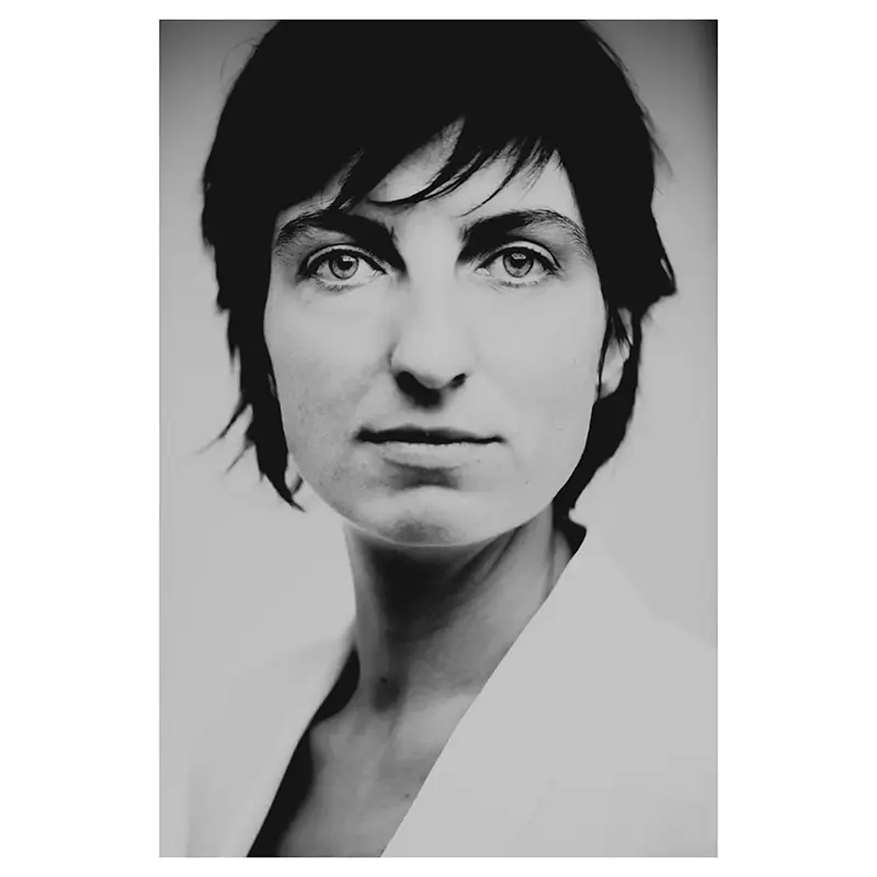 Portrait noir et blanc d’une femme aux cheveux courts bruns, gros plan trois-quarts, regard caméra, expression posée, lumière douce avec léger vignettage, veste claire type blazer par un photographe dans la drôme.