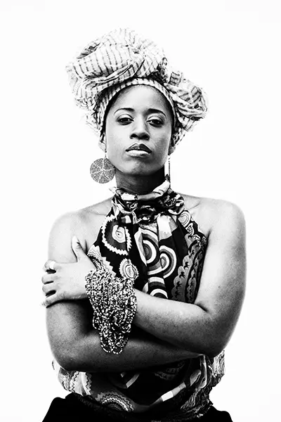 Portrait studio en noir et blanc high key d'une femme portant un turban volumineux et des bijoux, bras croisés sur fond blanc.