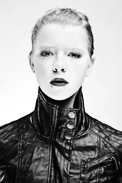 Portrait studio en noir et blanc high key d'une femme portant une veste en cuir noir à col montant sur fond blanc.