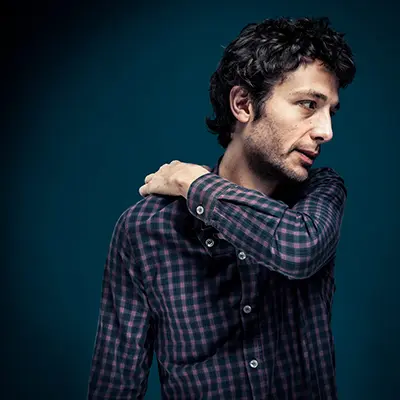 Portrait studio en couleur d'un homme en chemise à carreaux, la main sur l'épaule et le regard hors champ.
