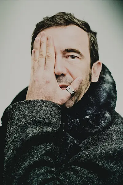 Portrait éditorial d'un homme masquant un œil avec sa main, portant un manteau en laine à col fourrure.