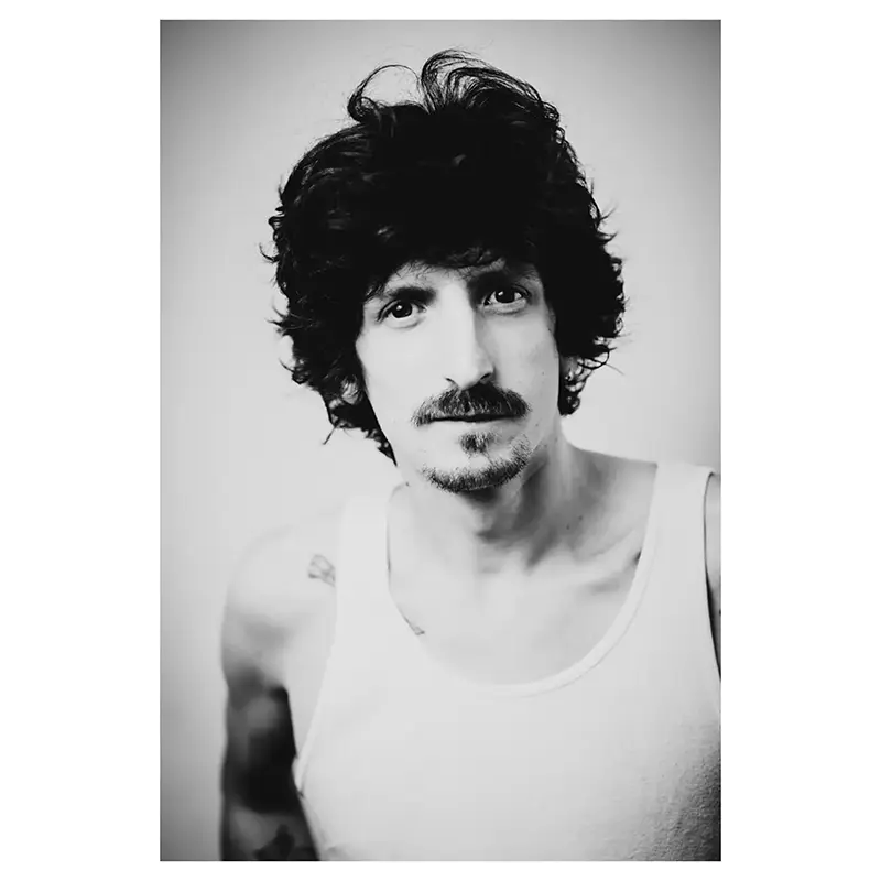 Portrait en studio photo en noir et blanc d’un homme jeune aux cheveux bouclés et moustache, plan poitrine trois-quarts, regard caméra, expression posée, lumière douce, débardeur blanc laissant voir de petits tatouages.
