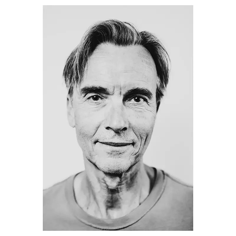 Portrait noir et blanc d’un homme mature, cheveux gris coiffés sur le côté, regard caméra et léger sourire, plan poitrine rapproché, lumière douce, t-shirt uni, par un photographe de portrait.