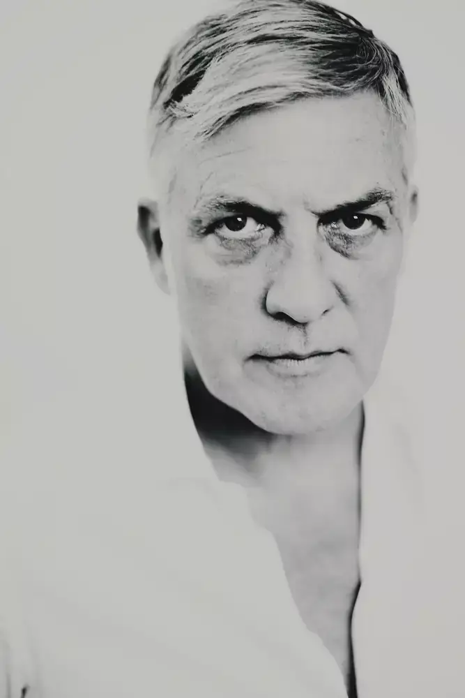 Gros plan d’un homme mature en chemise blanche, regard intense, rendu noir et blanc.